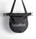 A black headfirst helmet bag with a wide adjustable webbing strap, plastic buckle closures, and a silver logo printed on the front, shown against a plain white background.