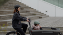 Woman riding a bike with a child in a stroller, both wearing helmets, on a city street. The Echo Pro - urban helmet by headfirst helmets is a matte black, modern, rounded bicycle helmet with small vents and a motion-sensing brake light, shown on a plain gray background.