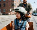Child wearing a helmet in a bike trailer with an adult behind, on a city street. headfirst helmets Echo Pro kids Green 